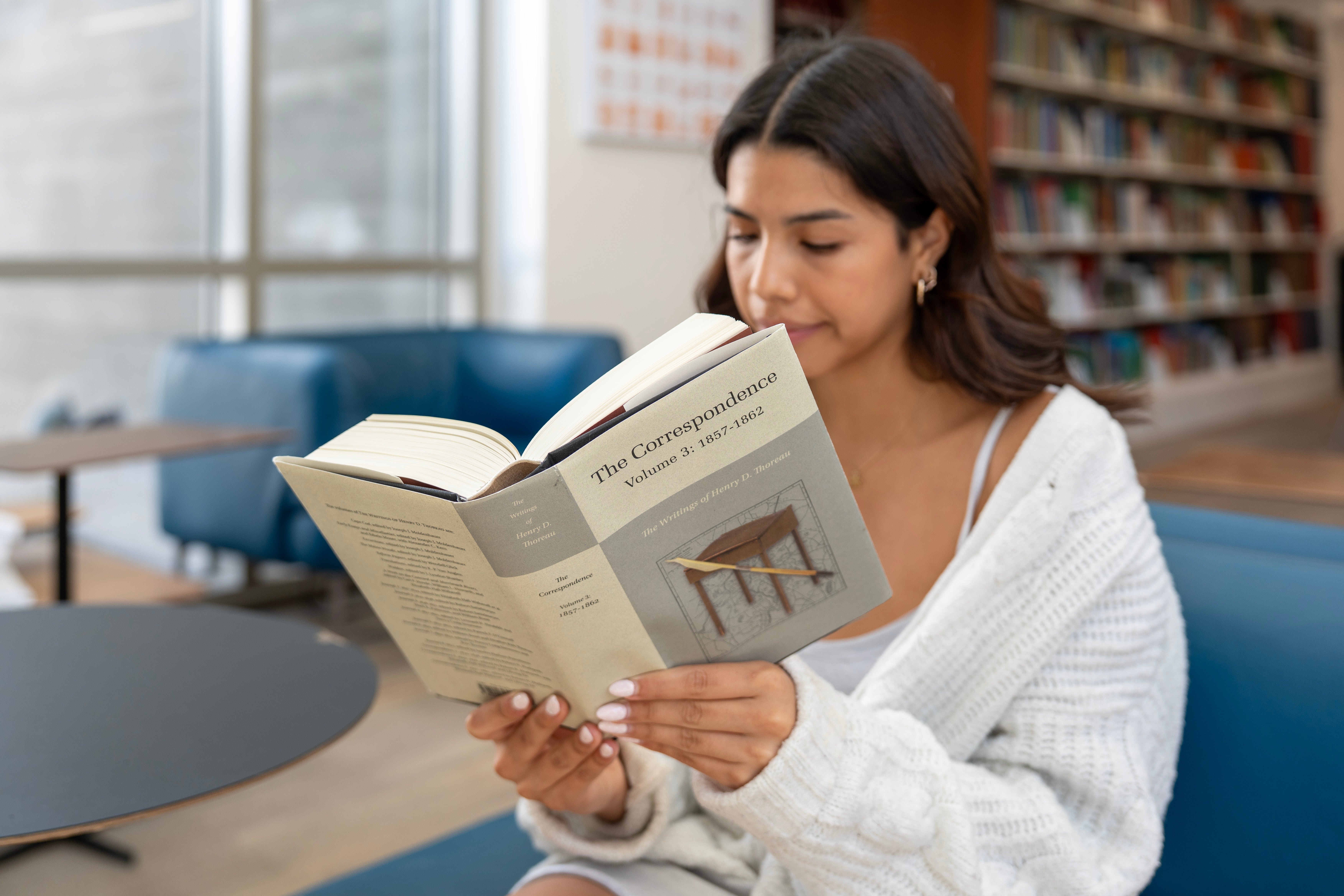 A UCSB student reads "Correspondence Volume 3." The student is seated on a blue lounge chair in the Sara Miller McCune Arts Library. A bookshelf is visible in the background.
